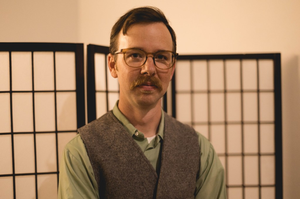 Portrait of Bill Kautz in a grey vest and green shirt, standing in front of a shoji screen