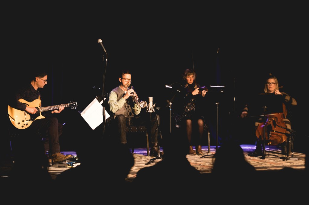Jeremy Drake on guitar, Bill Kautz on trumpet, Aria Peters on violin and Jessica Catron on cello performing on a darkened stage